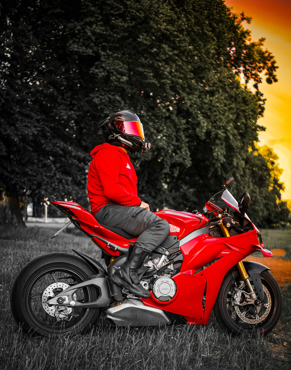 Motocyklista siedzący na czerwonym motocyklu sportowym przy drodze otoczonej drzewami.